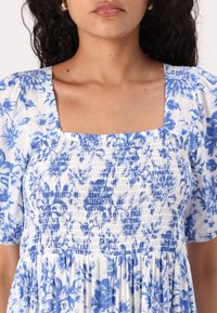 Robe fleurie bleue et blanche avec un décolleté carré, un corsage smocké et des manches courtes bouffantes, confectionnée dans un tissu léger.