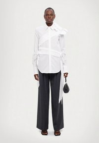 Camicia bianca con design asimmetrico e dettagli con nastri, abbinata a pantaloni larghi scuri e sandali neri, con una piccola borsa in mano.