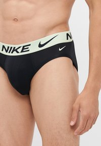 Sorte Nike-truser med en lys grønn midjebånd som har ordet "NIKE" i fet svart skrift. Mykt stoff og tettsittende design.