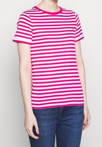 Camiseta a rayas rosa y blanca con mangas cortas, escote redondo y un pequeño logo en el pecho, combinada con jeans azules.