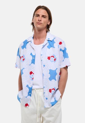 Homme aux cheveux bruns raides porte une chemise à manches courtes bleue et blanche à motif floral sur un t-shirt blanc et un pantalon blanc, les mains dans les poches.