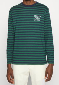 Langarmshirt mit Rundhalsausschnitt in Grün und Marineblau mit horizontalen Streifen, das auf der linken Brustseite weißen Text "ETUDES PARIS 75003" zeigt.