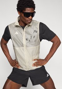 Ελαφρύ αμάνικο γιλέκο σε εκρού χρώμα με φερμουάρ μπροστά, μαύρες λεπτομέρειες και γραφικό τύπωμα. Συνδυάζεται με μαύρο κοντομάνικο T-shirt και σορτς.