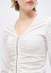 Femme portant un haut blanc froncé à manches longues avec une fermeture éclair argentée sur le devant et des boucles d'oreilles en forme d'anneau, fond neutre.