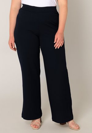 Vrouw draagt zwarte, hooggetailleerde, wijd uitlopende broek gecombineerd met witte, sandaalachtige sandalen met hak, staand tegen een effen achtergrond.