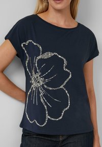 Marineblauw T-shirt met een pailletten bloemenontwerp, ronde halslijn, korte mouwen en een relaxte pasvorm, gecombineerd met een donkere spijkerbroek.