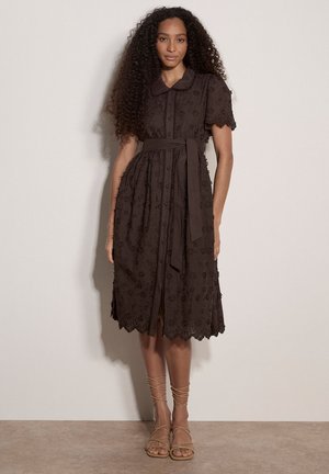 Kobieta z długimi kręconymi włosami, ubrana w ciemnobrązową sukienkę do kolan z kwiatowym haftem, krótkimi rękawami, zapinana na guziki z przodu i zawiązanym paskiem w talii, stojąca.