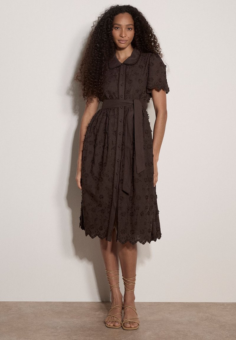 Femme aux longs cheveux bouclés portant une robe marron foncé longueur genoux avec broderie florale, manches courtes, boutonnée devant et ceinture nouée à la taille, debout.