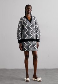 Black and white zigzag patterned sweater dress with a V-neck, ribbed cuffs, and hemline. Worn with white bow-accented sandals.