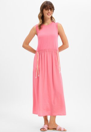 Frau in einem ärmellosen, pinken Midikleid mit Taillen-Kordelzug, trägt Sonnenbrille und pinke Sandalen, steht mit den Händen hinter dem Rücken.