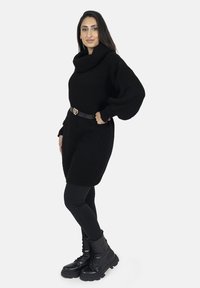 Robe pull en maille noire avec un col drapé et des manches longues, cintrée à la taille par une ceinture noire ornée d'une boucle en forme de cœur dorée. Assortie avec des leggings noirs et des bottes de combat noires à semelles épaisses.