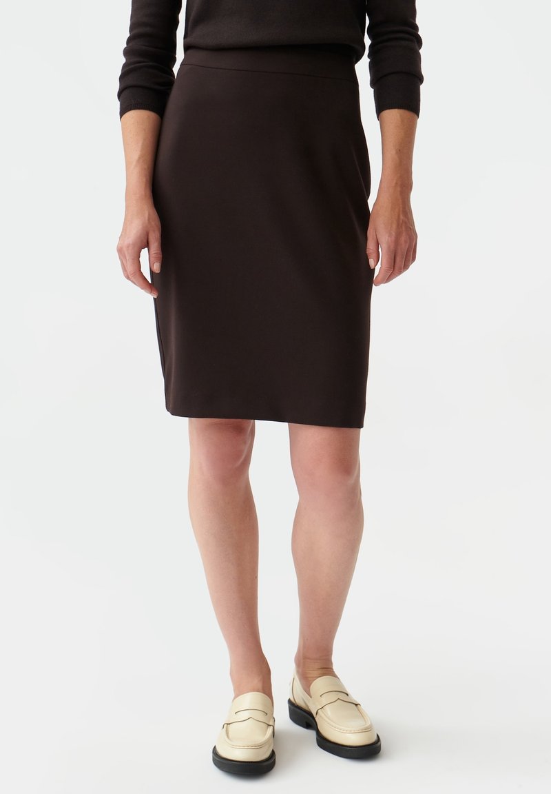 Femme portant une jupe noire longueur genou et des mocassins beige, debout devant un fond blanc uni.