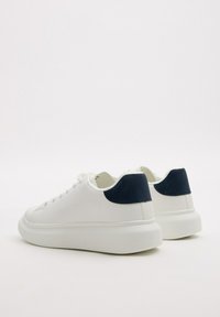 Zapatillas blancas con parte superior de cuero liso, detalles en azul marino en el talón, suelas gruesas de goma blanca y diseño minimalista, con cordones redondos.