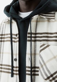 Camicia a quadri in misto lana bianca, marrone e beige, indossata sopra un felpa verde scuro con cappuccio e coulisse, dotata di una tasca frontale.