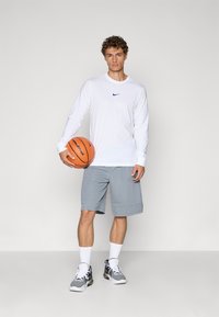 Hvid langærmet skjorte med et blåt Nike-logo, grå shorts og sorte sportsko. Holder en orange basketball, står foran en neutral baggrund.