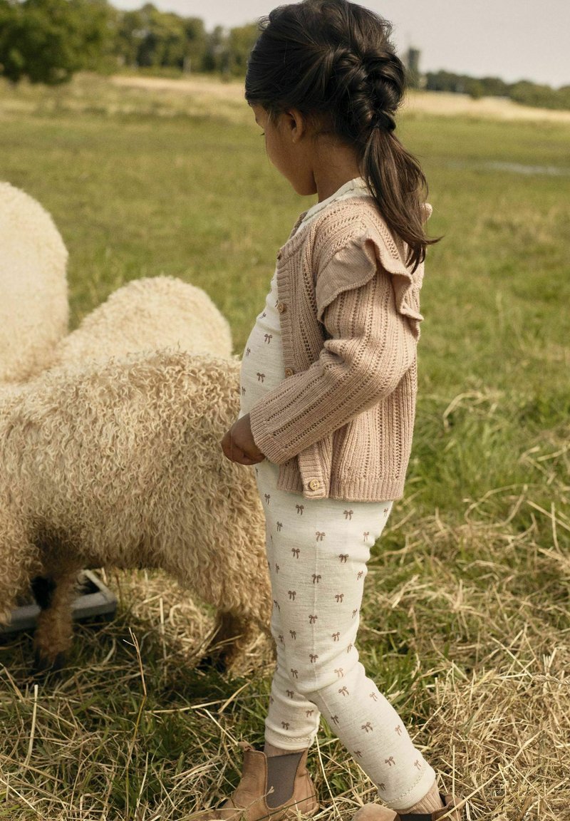 Un enfant portant un cardigan en tricot beige, un pantalon crème à motifs marron et des bottines brunes près de moutons duveteux dans un champ herbeux.