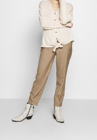 Beige långärmad topp med knappar och knutdetalj i midjan, kombinerad med ljusbruna smalrandiga byxor och vita ankelstövlar i ormskinnsmönster.