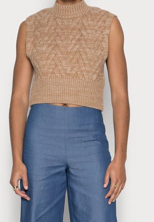 Top court sans manches beige en maille avec col roulé et motif zigzag, associé à un pantalon large bleu taille haute, porté par une personne.