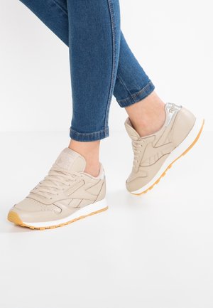 Persoon draagt beige sneakers met witte middenzolen en rubberen buitenzolen in kauwgomkleur, gecombineerd met strakke blauwe spijkerbroeken, staand op een wit oppervlak.