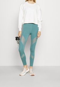 Sudadera corta de color gris claro con un logo sutil; mallas en color teal con paneles de malla geométricos; zapatillas deportivas en color crema y coral; botella de agua en la mano.