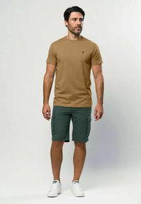 Brūna sporta krekla ar apaļu kakla izgriezumu kombinācija ar zaļiem cargo īsiem šortiem, kam ir sānu kabatas. Pabeigts ar baltām kedām. Vienkāršs, ikdienišķs izskats.