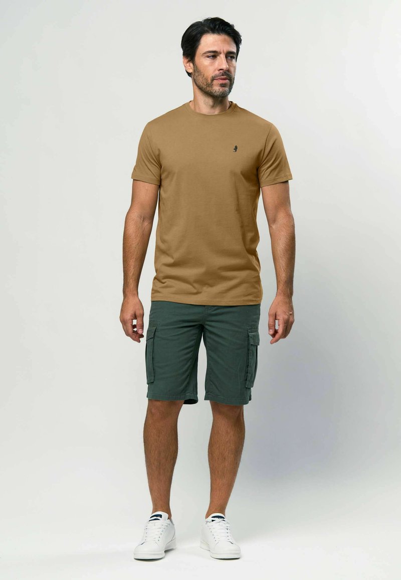 Brūna sporta krekla ar apaļu kakla izgriezumu kombinācija ar zaļiem cargo īsiem šortiem, kam ir sānu kabatas. Pabeigts ar baltām kedām. Vienkāršs, ikdienišķs izskats.