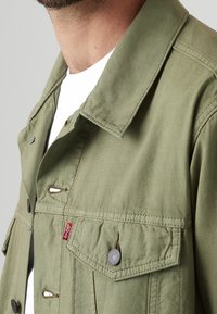 Chaqueta verde caqui hecha de un tejido duradero, con un cuello puntiagudo, múltiples bolsillos en la parte delantera y botones a presión de metal.