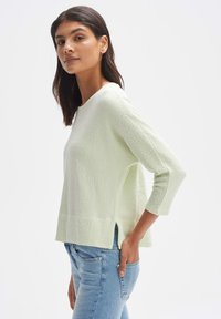 Hellgrüner, strukturierter Strickpullover mit 3/4-Ärmeln, Boot-Ausschnitt und seitlichen Schlitzen, kombiniert mit blauen Jeans.