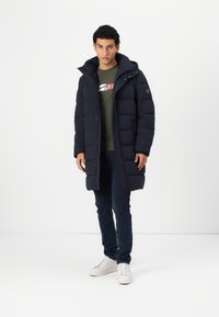 Jūras spēku puffer jakas ar kapuci, ar quilted dizainu un priekšējiem pogām. Valkāta virs zaļās T-krekla un tumšajām džinsiem, apvienota ar baltajiem sporta apaviem.