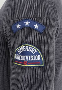 Grauer Strickpullover mit marineblauen Stoffakzenten. Enthält Patches mit weißem Text und Sternen in Blau-, Gelb- und Rottönen.