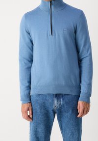 Pull bleu clair à fermeture éclair avec col montant, poignets et ourlet à côtes. Présente un petit patch logo sur la poitrine. Texture en maille douce.