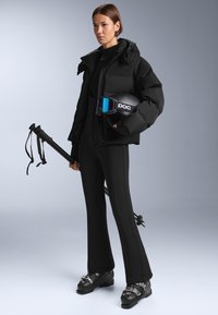 Černá prošívaná bunda s kapucí, přiléhavý černý lyžařský oblek, černé lyžařské boty s kovovými přezkami, lyžařské hole a helma s modře tónovaným štítem.