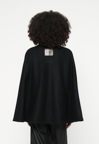 Μαύρη μάλλινη κάπα με λεία υφή, διαθέτει κεντρική ραφή στην πλάτη και ετικέτα στο λαιμό. Κρέμεται χαλαρά, φτάνοντας μέχρι το μέσο της πλάτης.
