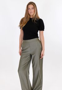 Młoda kobieta o długich blond włosach, ubrana w czarną bluzkę z krótkim rękawem i luźne oliwkowozielone spodnie, stojąca na białym tle.