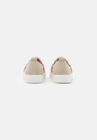 Beige Leinensneaker mit weißen Gummisohlen, die eine strukturierte Oberfläche und ein minimalistisches schnürsenkelloses Design aufweisen, von hinten gesehen.