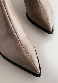Bottines à bout pointu en cuir taupe clair avec une finition texturée et une semelle en caoutchouc noir. Des coutures visibles ajoutent des détails sur les côtés.