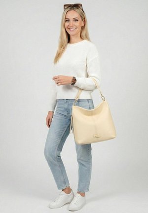 Blonde Frau trägt weißen Pullover, helle Jeans, weiße Sneaker, Sonnenbrille auf dem Kopf, hält eine beiger Handtasche und lächelt vor schlichtem Hintergrund.