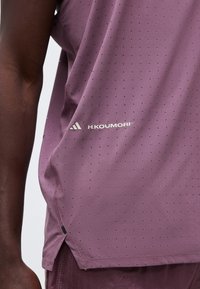 Ramię i tułów osoby w fioletowej sportowej koszulce i szortach z małymi czarnymi kropkami oraz logo "H.KOUMORI" na koszulce.