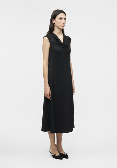 Γυναίκα στέκεται φορώντας μαύρο αμάνικο μίντι φόρεμα με ντραπέ ντεκολτέ και μαύρες γόβες με λουράκι στη φτέρνα και μυτερή μύτη σε λευκό φόντο.