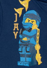 Maglietta grafica blu con un personaggio ninja Lego che tiene una catena e shuriken, su uno sfondo blu scuro, con lettere dorate che spellano "JAY."