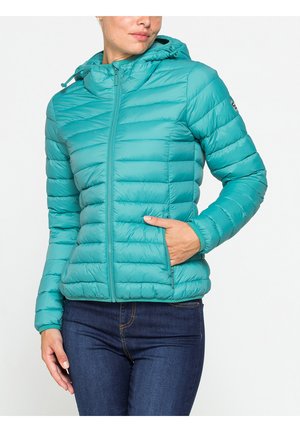 Donna che indossa una giacca trapuntata verde teal con cappuccio, zip frontale e tasche laterali, abbinata a jeans blu scuro.