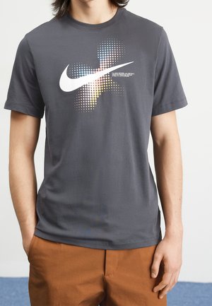 Mann trägt ein dunkelgraues Nike T-Shirt mit einem bunten, gepunkteten Swoosh-Logo und braunen Hosen, steht mit einer Hand in der Tasche.