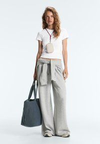 Femme portant un t-shirt blanc, un pantalon de survêtement gris ample avec un sweat assorti noué à la taille, portant un sac fourre-tout bleu foncé et un collier pochette beige.
