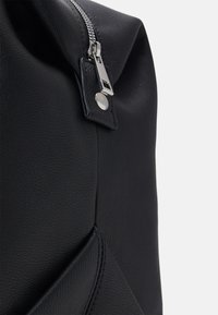 Sort tekstureret læderrygsæk hjørne, med en sølvfarvet lynlås og hardware, samt en syning og glat overflade.