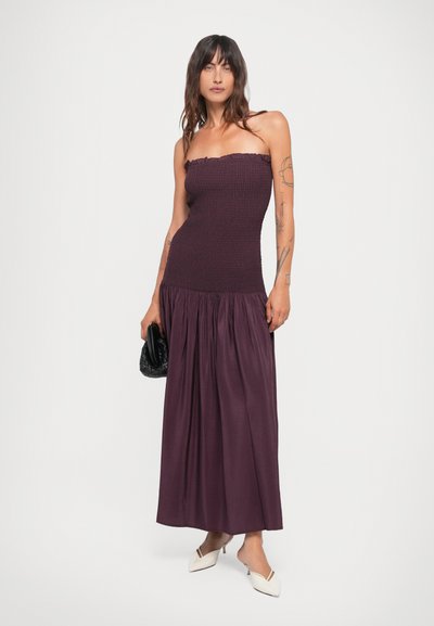 Lange, strapless jurk van donkerpaarse stof met een gerimpelde lijfje en een vloeiende, geplooide rok. Gecombineerd met een zwarte clutch en witte muiltjes.