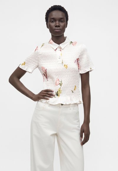Femme portant un chemisier smocké blanc à fleurs, manches courtes avec col et un pantalon blanc taille haute, posant avec une main sur la hanche.