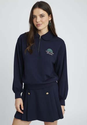 Sweatshirt zippé bleu marine avec col et graphique vert brodé, associé à une jupe plissée bleu marine avec des accents de boutons dorés.