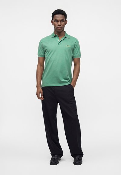 Homme debout portant un polo vert à manches courtes, un pantalon large noir et des chaussures noires à pompons sur un fond uni.