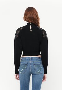 Pull cropped noir à manches longues, avec des accents en dentelle sur les épaules, un décolleté en goutte dans le dos avec un embellissement en métal, et une texture lisse.