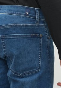 Siniset farkut, joilla on rakenteellinen istuvuus, takatasku, sisäsaumapistot ja merkki, jossa lukee "kaikille ihmisille".